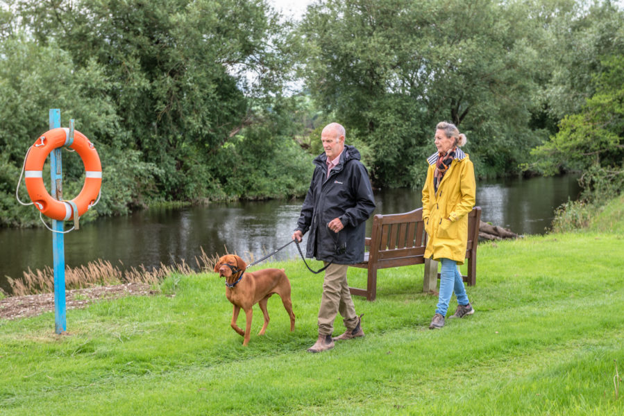 A man in a black coat and green trousers and a woman wearing a yellow mac and jeans walking their Vizsla dog with the river in the background and a lifeguard ring