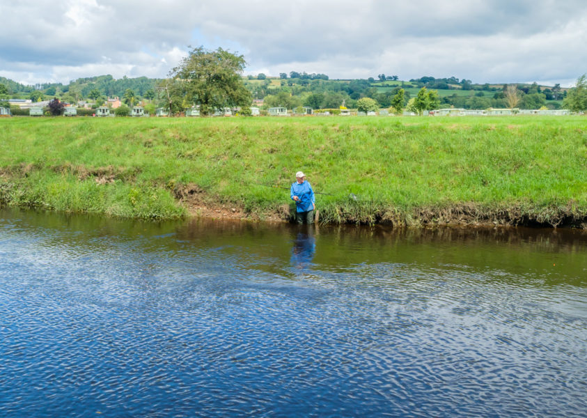 A man wearing waterproofs standing in the river, fishing. In the background is a river bank and caravans.