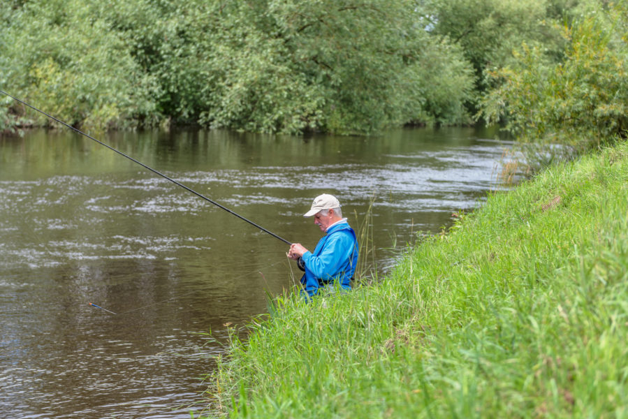 A man with a khaki cap and bright blue mac fishing on a river bank
